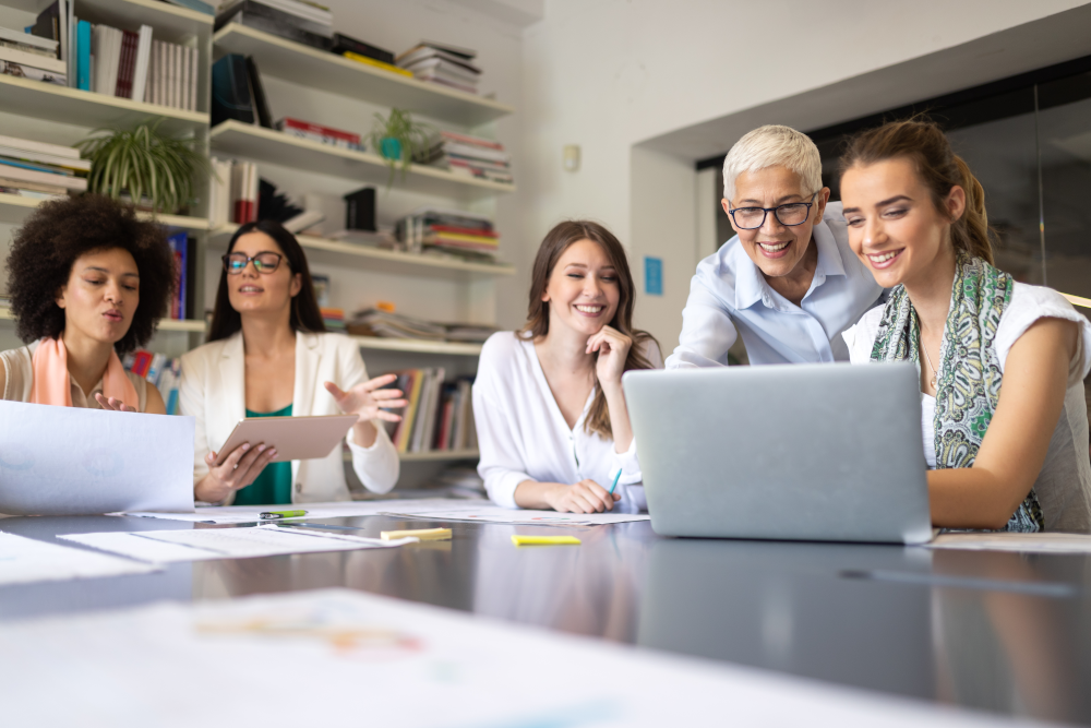 An office meeting depicting multigenerational harmony and productivity. A diverse group of employees from different generations are seated around a large conference table, smiling and engaged in a constructive discussion.