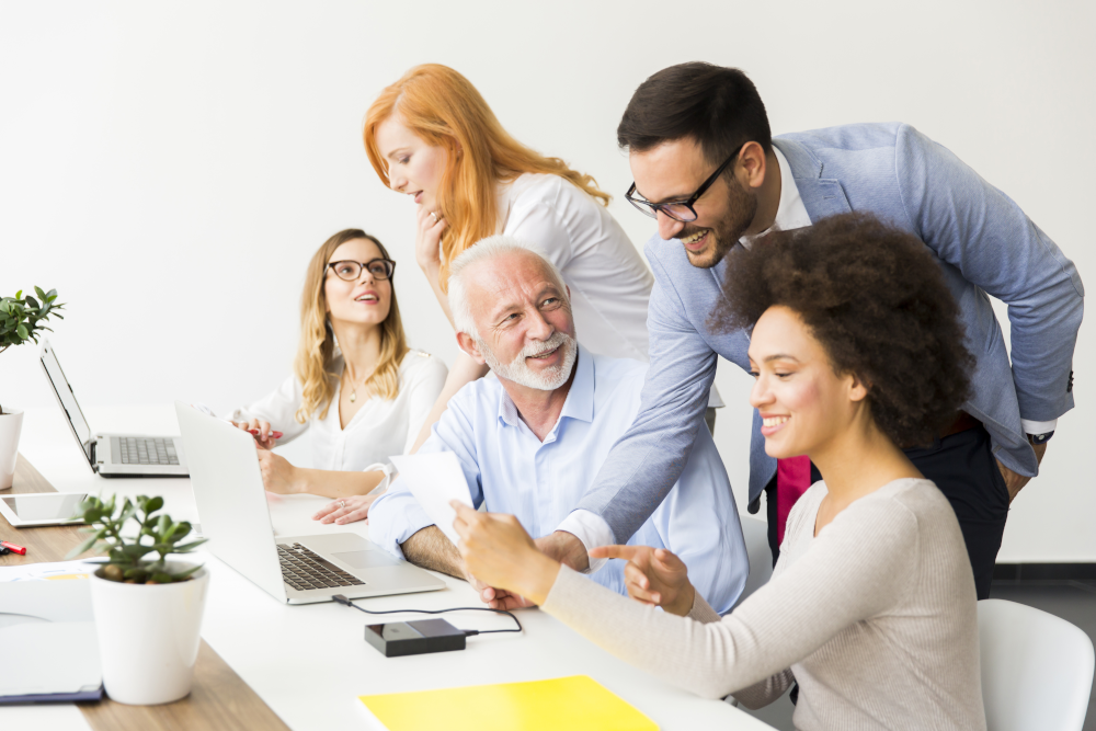 An office scene depicting a multi-generational workplace with employees of various ages collaborating around a conference table.